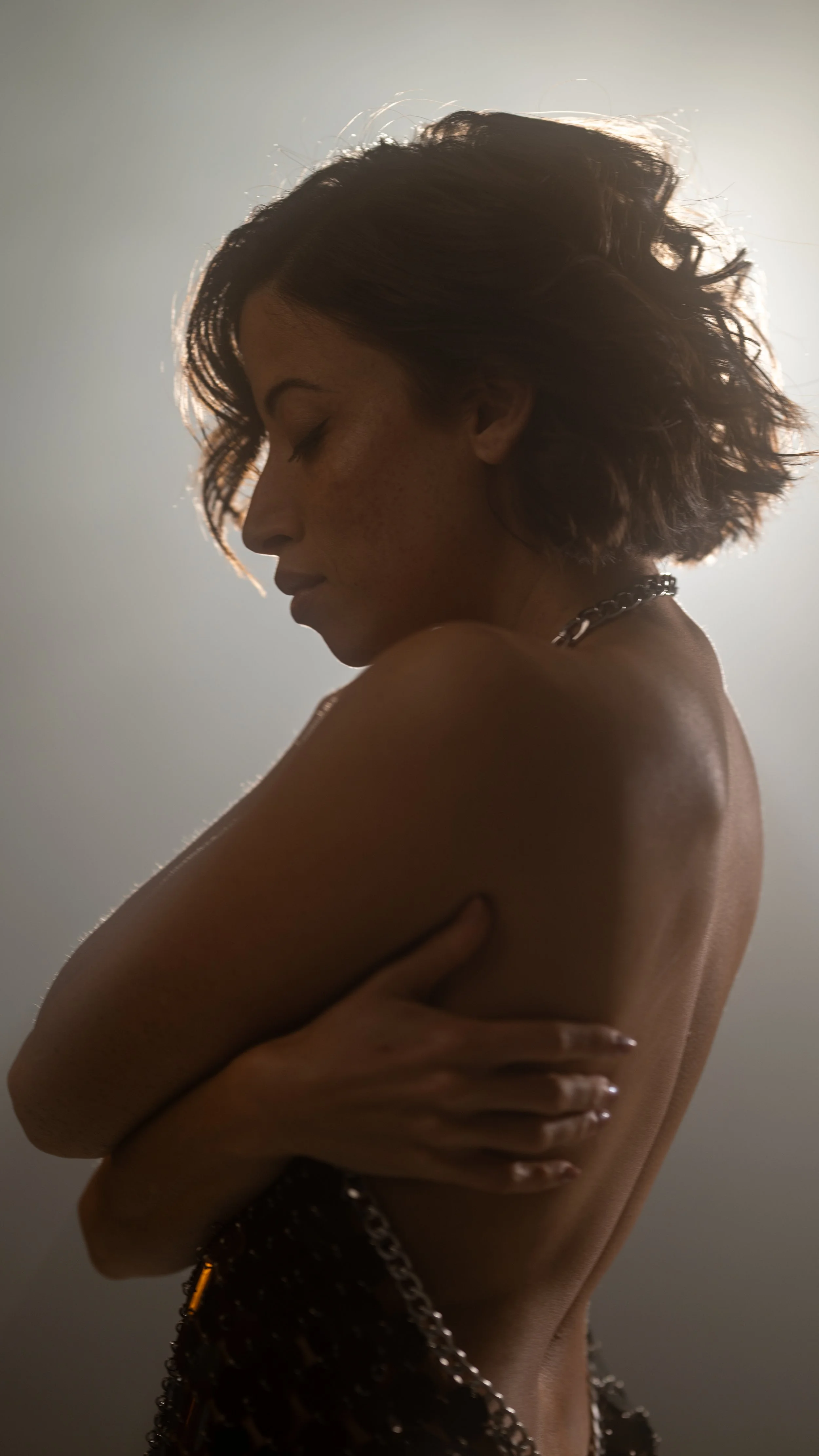 Porträt einer Frau mit geschlossenen Augen, umarmt sich selbst, im Profil sichtbar, vor neutralem Hintergrund.