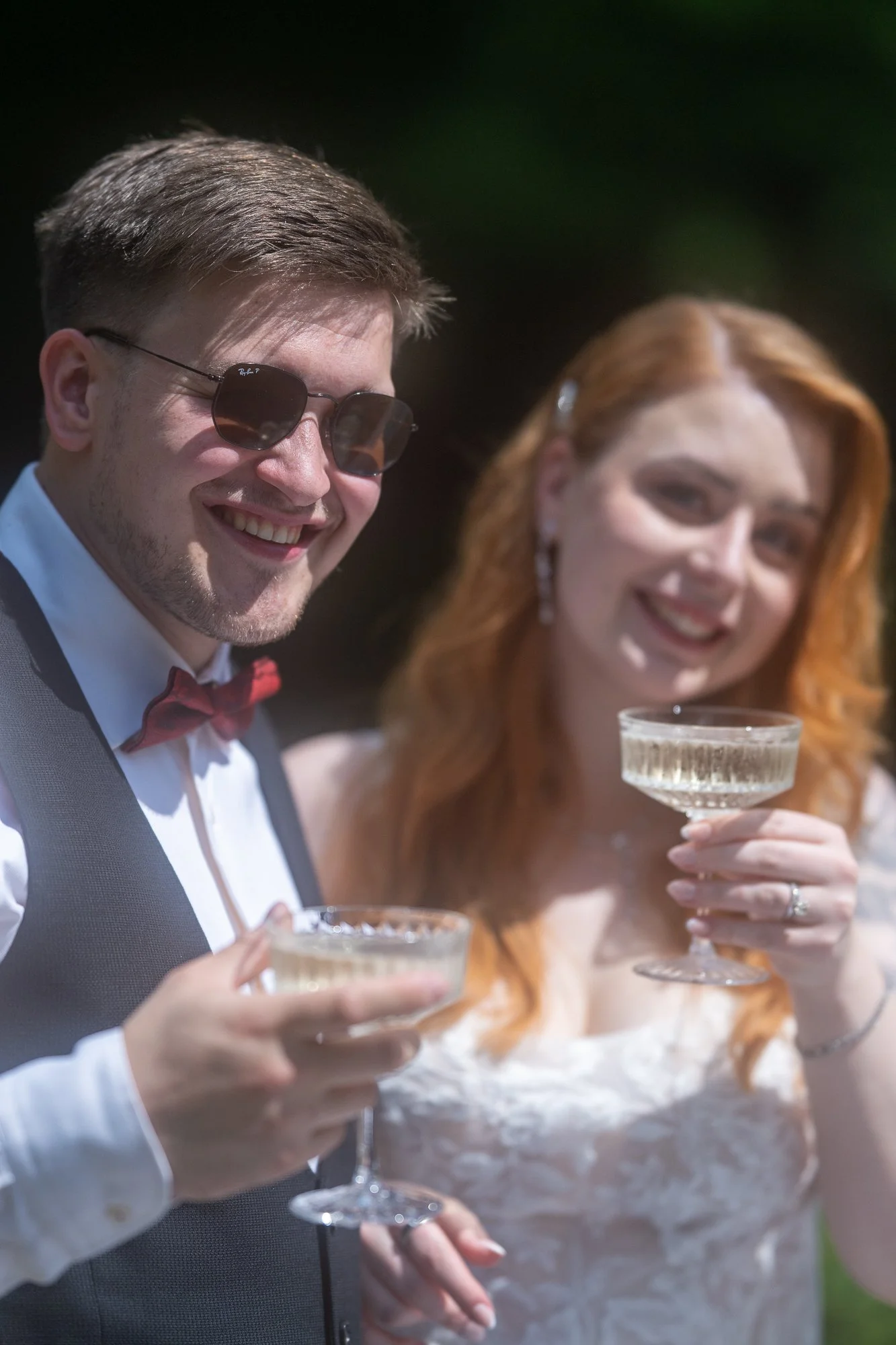 Ein Mann trägt eine schwarze Sonnenbrille und einen Smoking mit roter Fliege, während eine Frau mit roten Haaren in einem weißen Kleid ein Champagnerglas hält. Sie trinken Champagner und lächeln.