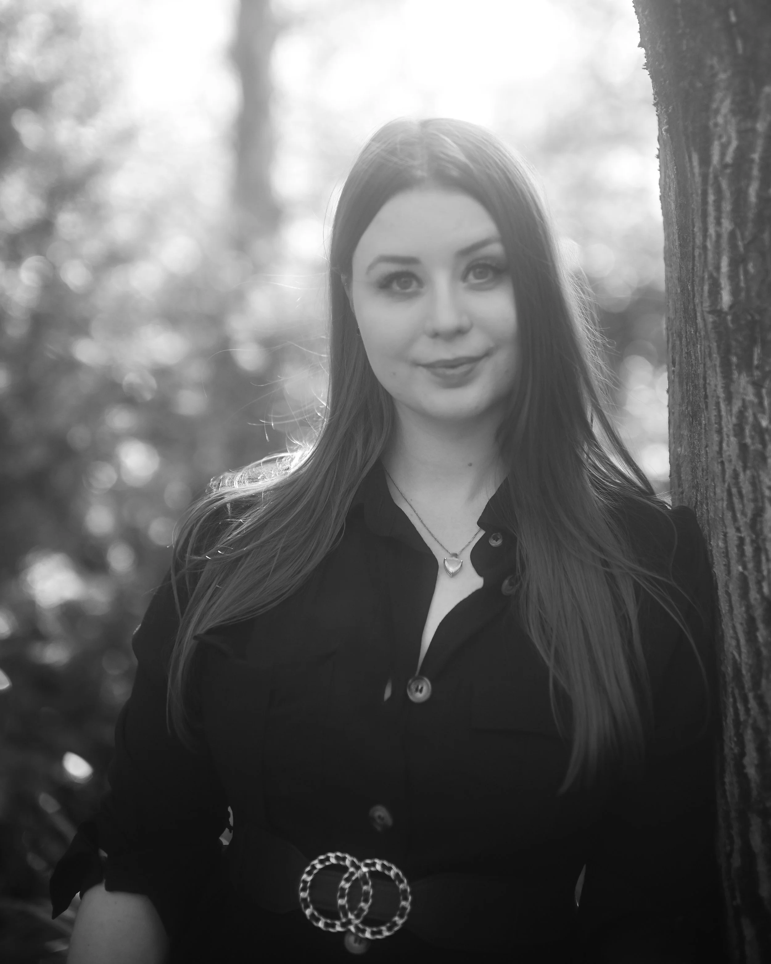 Schwarzes und weißes Portrait einer jungen Frau mit langen Haaren, die vor einem Baum steht, mit Blumen im Hintergrund, schwarzer Bluse und Herzanhänger, graue Kette, im Freien bei Tageslicht.