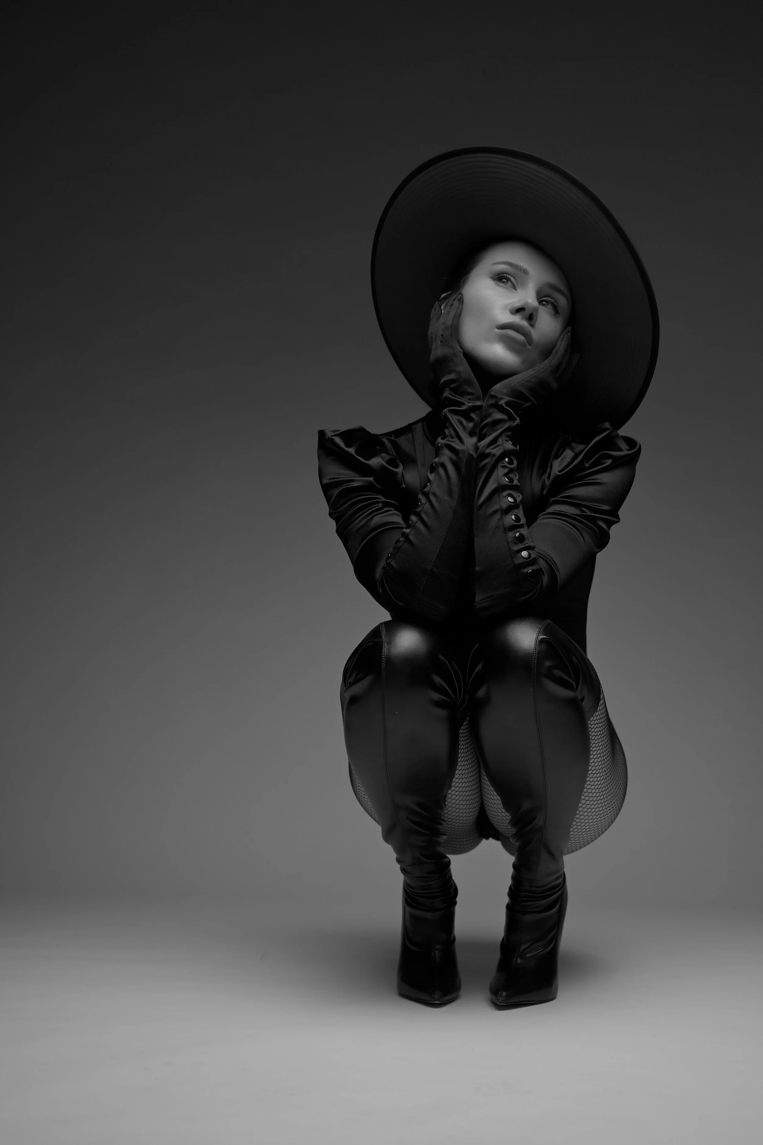 Schwarz-weiße Fotografie einer Frau mit großem Hut, die ihre Hände an das Gesicht legt, sitzt auf den Fersen, trägt ein elegantes, glänzendes Outfit und hat einen nachdenklichen Ausdruck.