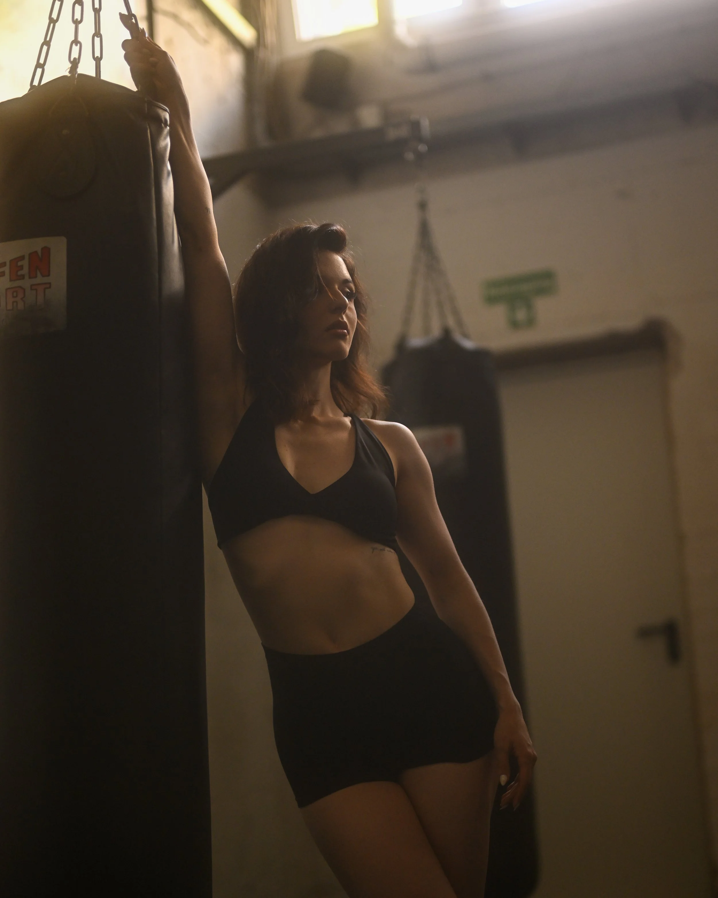 Eine junge Frau in sportlicher Kleidung steht in einem Raum mit minimaler Beleuchtung. Sie hat braune, lockige Haare und wirkt nachdenklich. Es sind Boxsäcke im Hintergrund sichtbar.