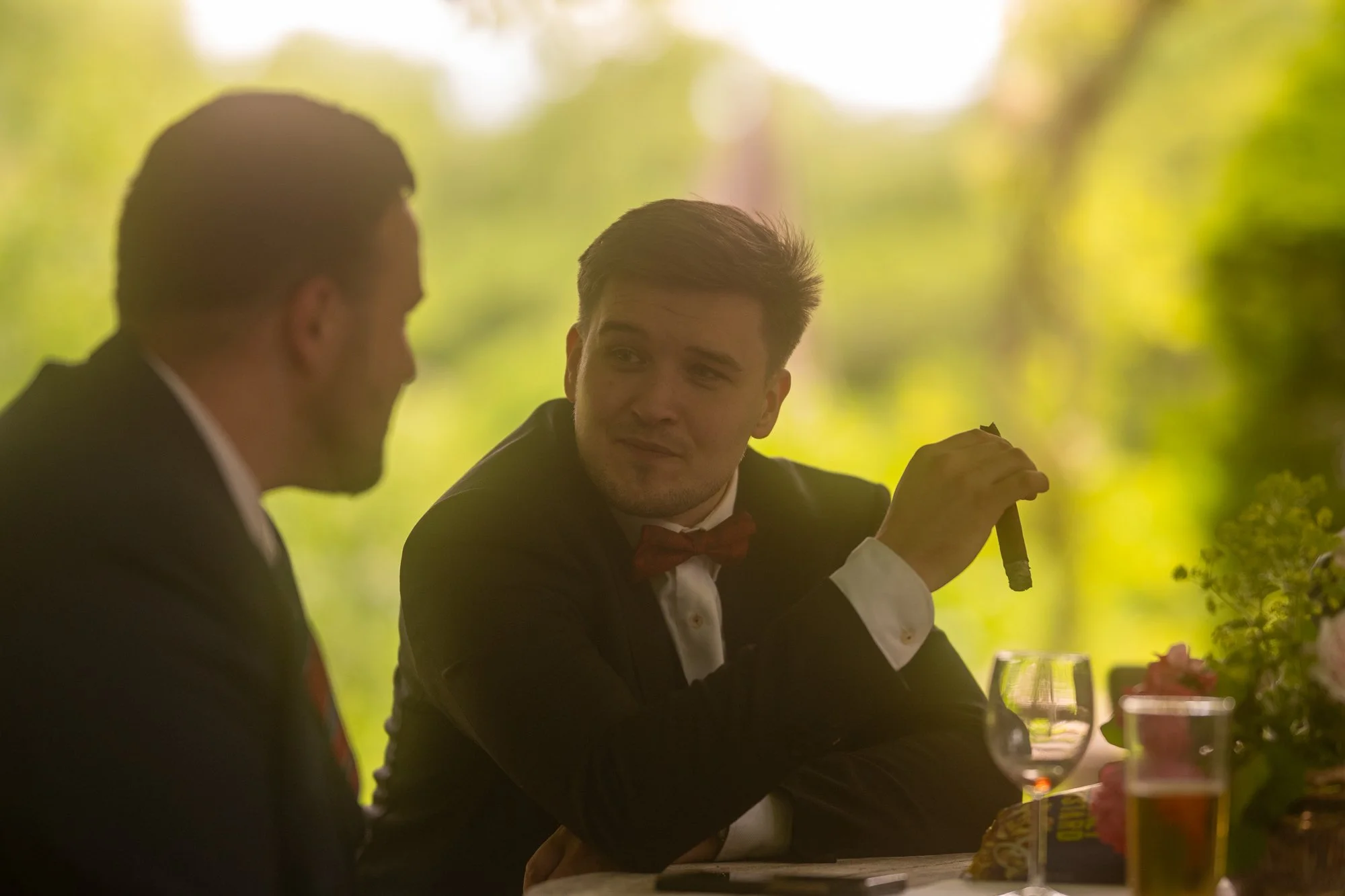 Zwei Männer in formeller Kleidung, einer mit Zigarette, unterhalten sich in einem gehobenen Restaurant im Freien mit grüner Umgebung.
