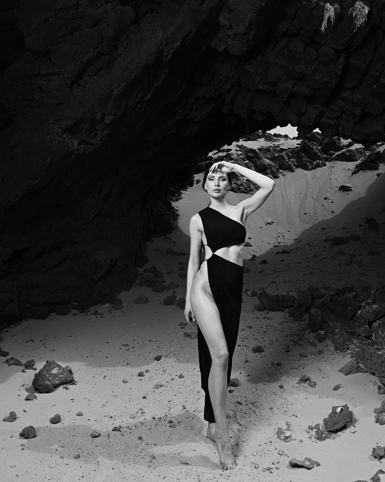 Eine schwarz-weiße Fotografie einer jungen Frau in einem langen schwarzen Kleid, die barfuß an einem Strand steht, umgeben von Felsen und Sand, unter einem großen Felsbogen.