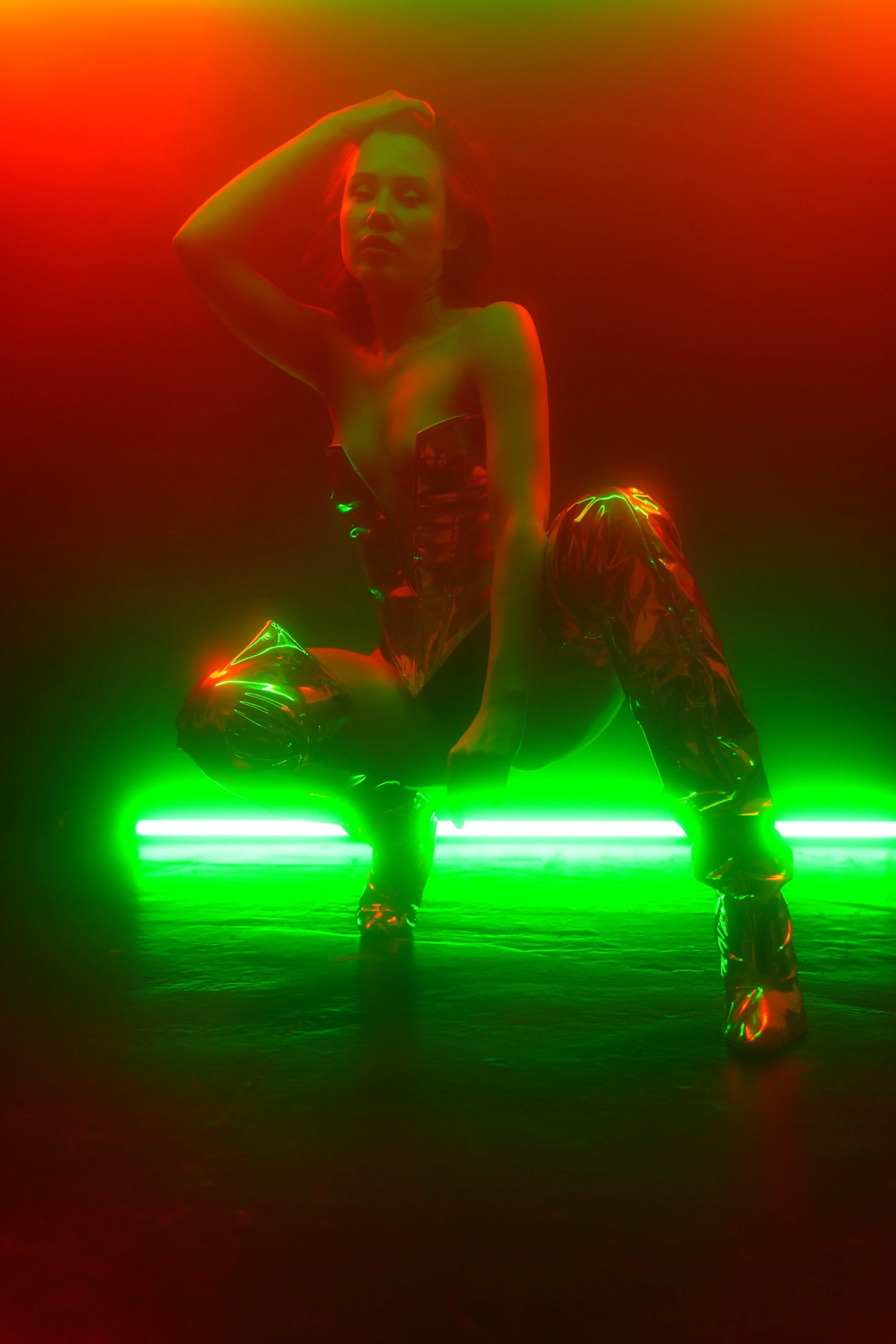 Ein Model sitzt auf dem Boden vor einer Neon-Lichtinstallation in Rot und Grün, trägt glänzende, metallische Kleidung und High Heels.