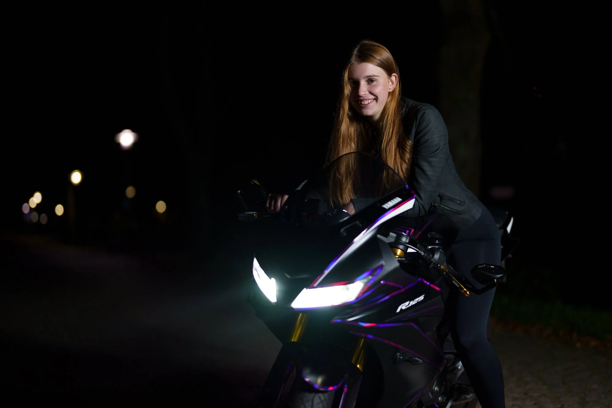 Junge Frau mit langen roten Haaren sitzt auf einem schwarzen Motorrad bei Nacht und lächelt.