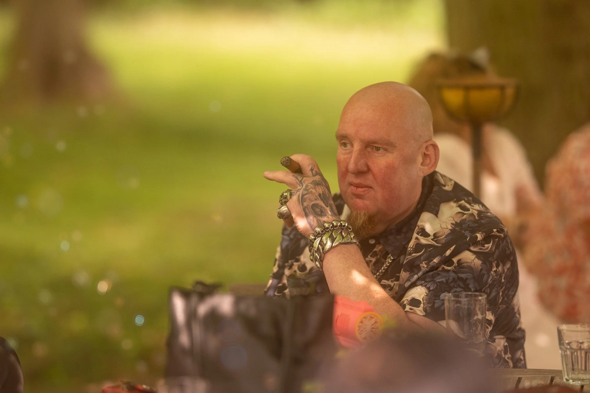 Mann mit Bart und Tattoos, trägt eine Lounge-Hose, sitzt an einem Tisch in einer Bar oder einem Restaurant, hält eine Zigarre in der Hand, im Hintergrund Frauen und eine warme Beleuchtung.