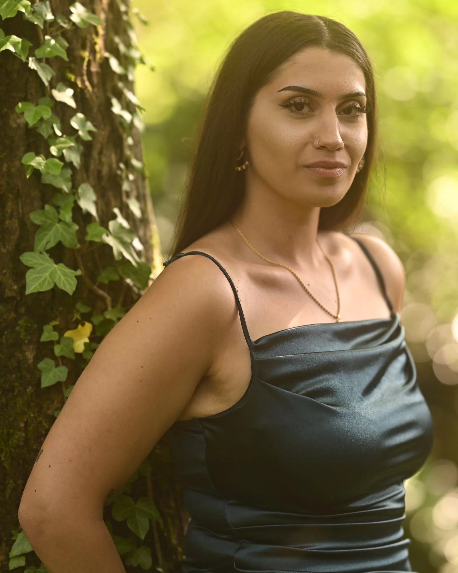 Eine junge Frau steht vor einem Baum mit Efeu, trägt ein schwarzes Kleid und Goldschmuck, im natürlichen Licht überrascht.