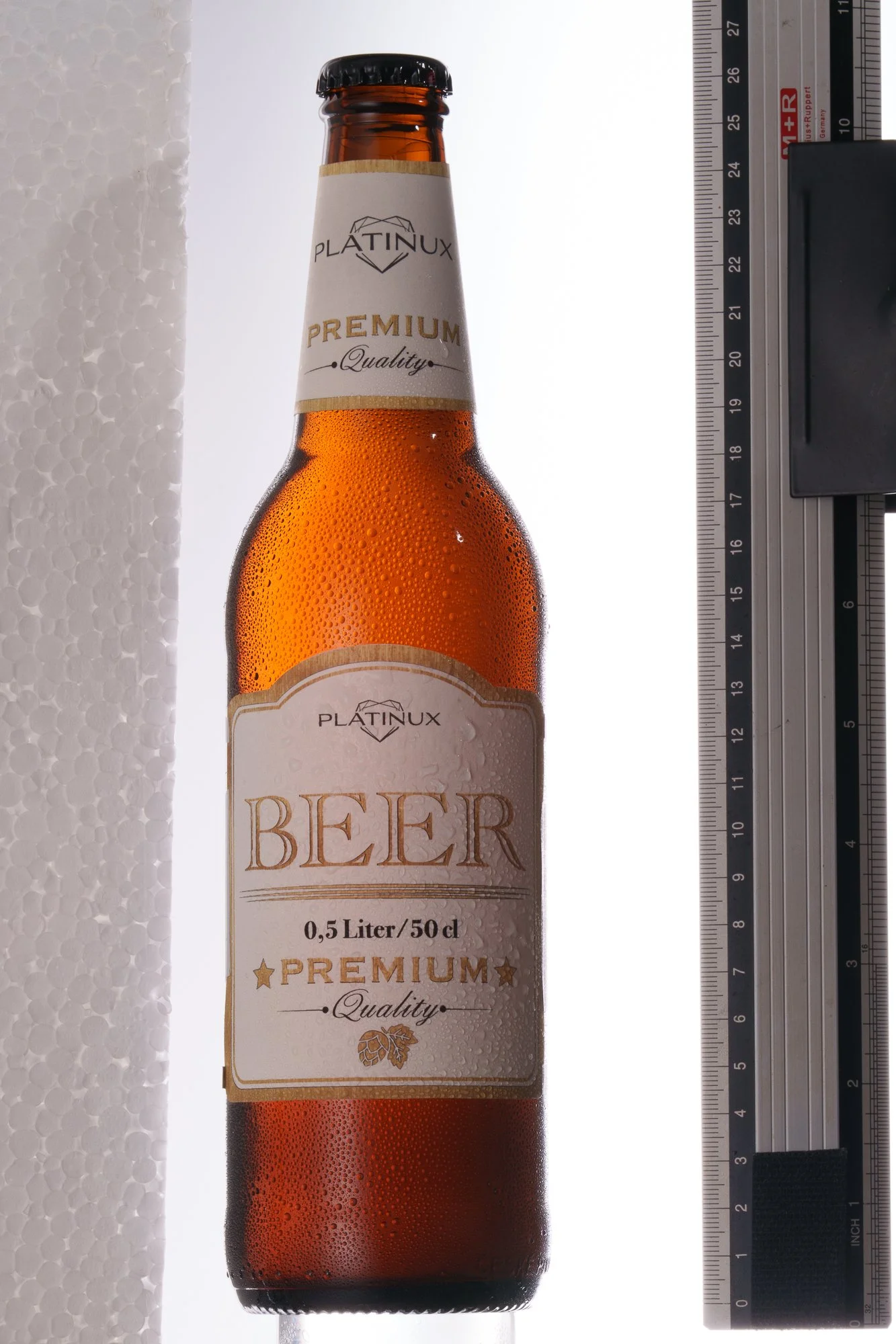 Braune Bierflasche mit Wassertröpfchen, weißes Etikett mit goldenen und schwarzen Schriftzug, daneben ein Lineal zur Größenmessung.