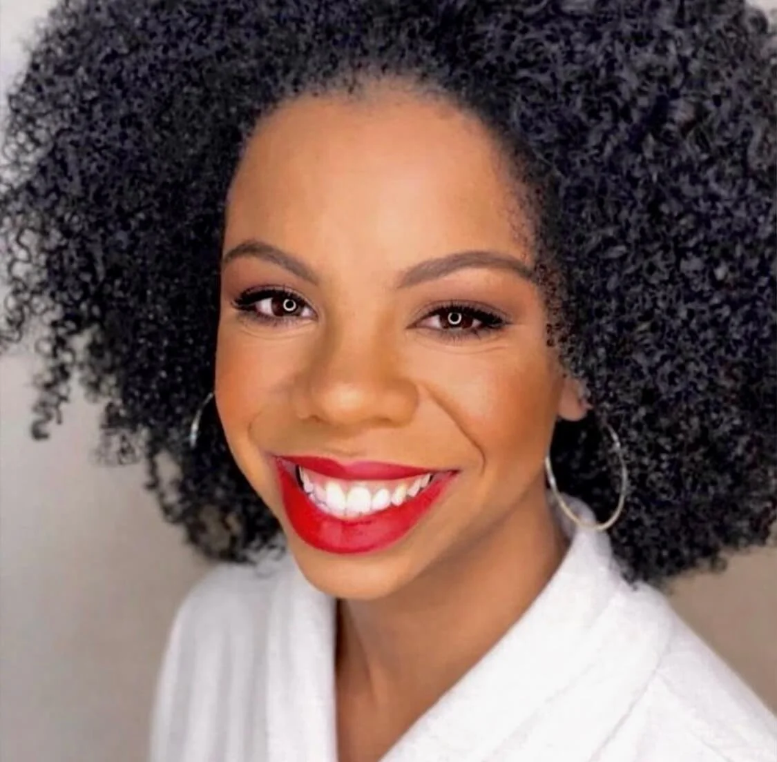 Bold red lip event makeup black skin by Lara Claudy, PhiladelphiaA woman with medium skin, curly black hair, red lipstick, and hoop earrings smiling at the camera.