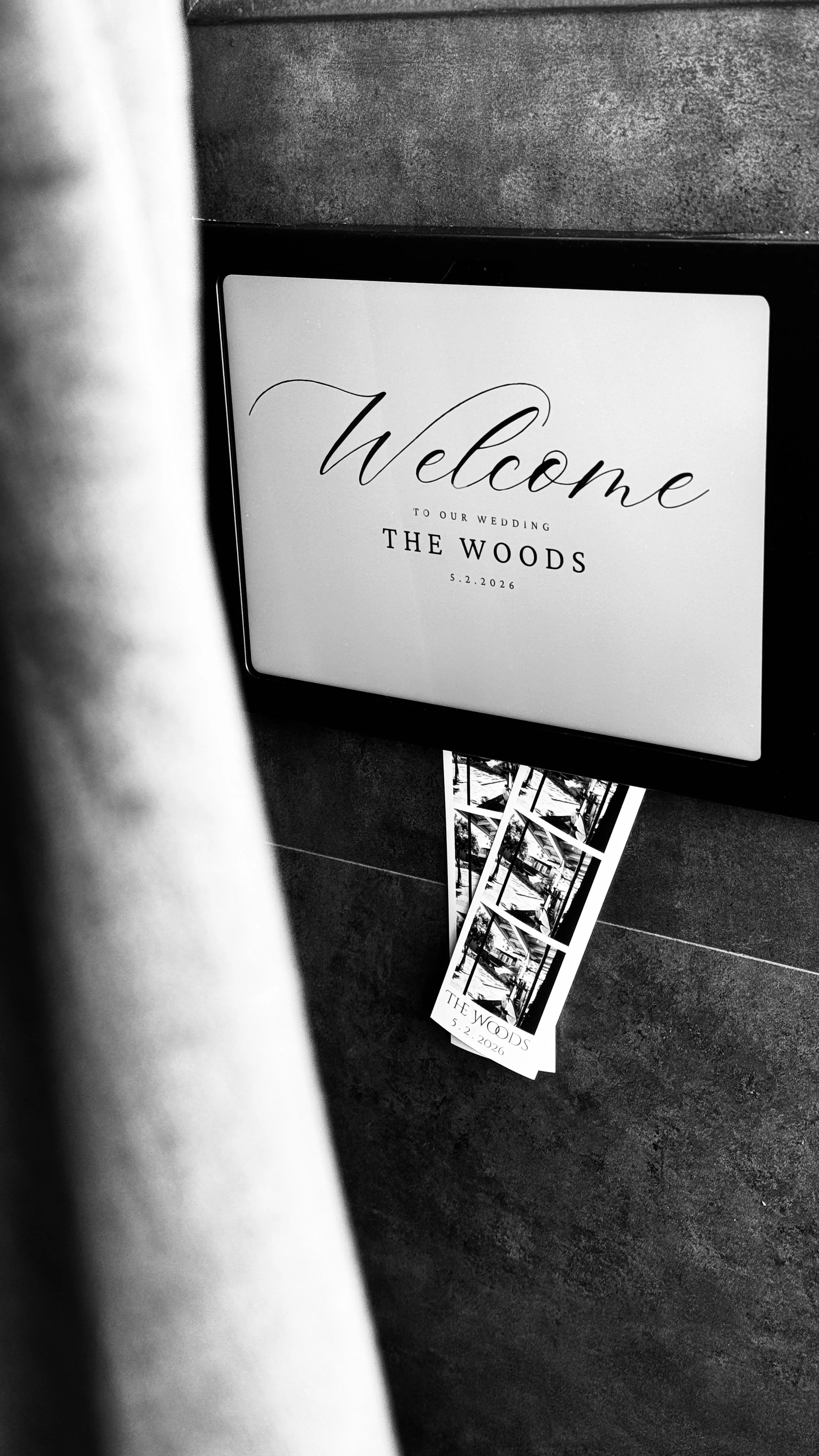 A digital sign displaying a wedding welcome message for 'The Woods' on May 2, 2026, with 2" x 6" photo strips on a slate stone surface.
