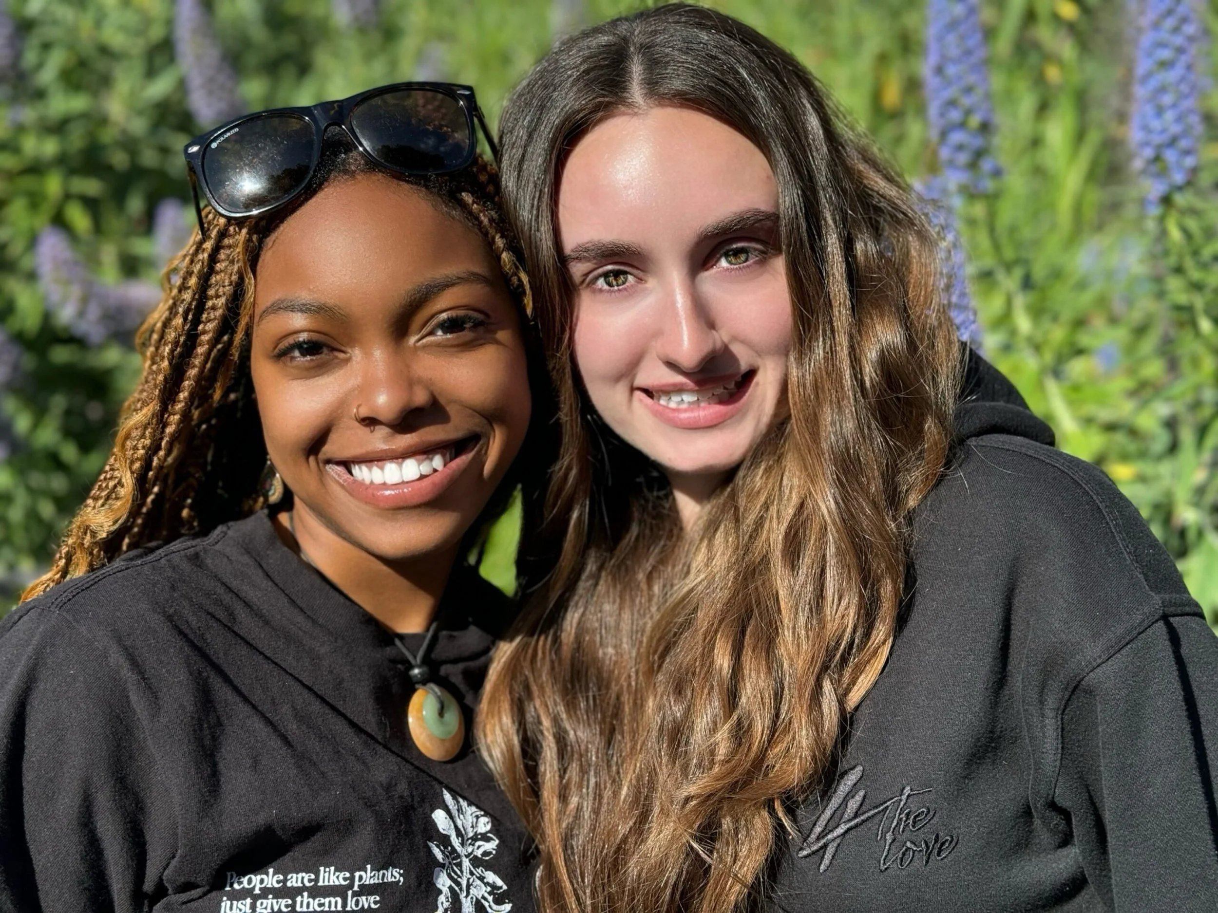 Two women smiling close to each other, one with dark skin and braided hair wearing sunglasses on her head, and the other with light skin and long wavy hair. They are outdoors with a blurry background of greenery and purple flowers.
