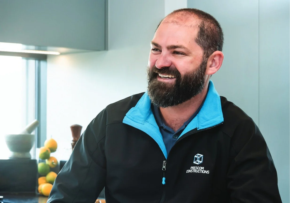 Smiling man with a beard wearing a black jacket and blue shirt, sitting indoors near a window with fruit and bowls in the background.