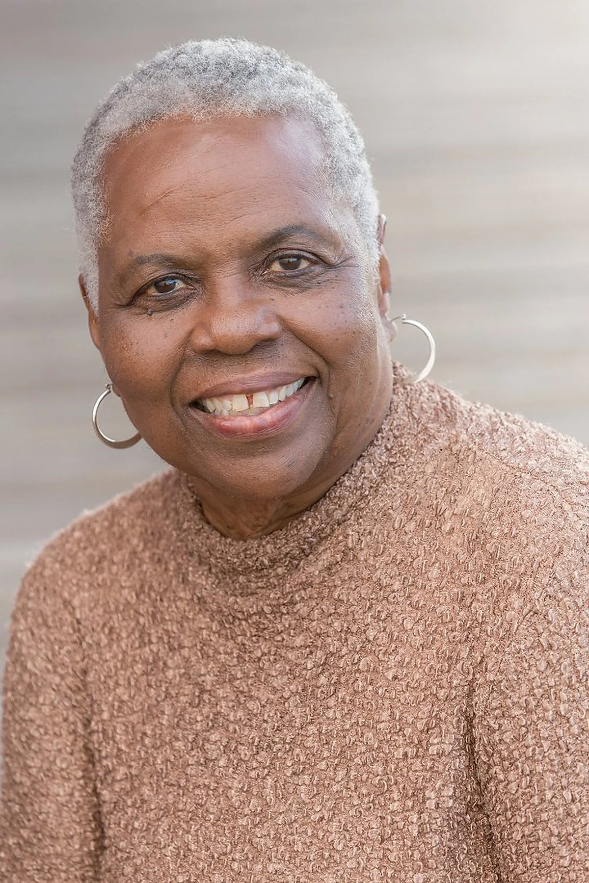 Portrait of an elderly woman with short, gray hair smiling, wearing hoop earrings and a beige textured sweater against a blurred background.