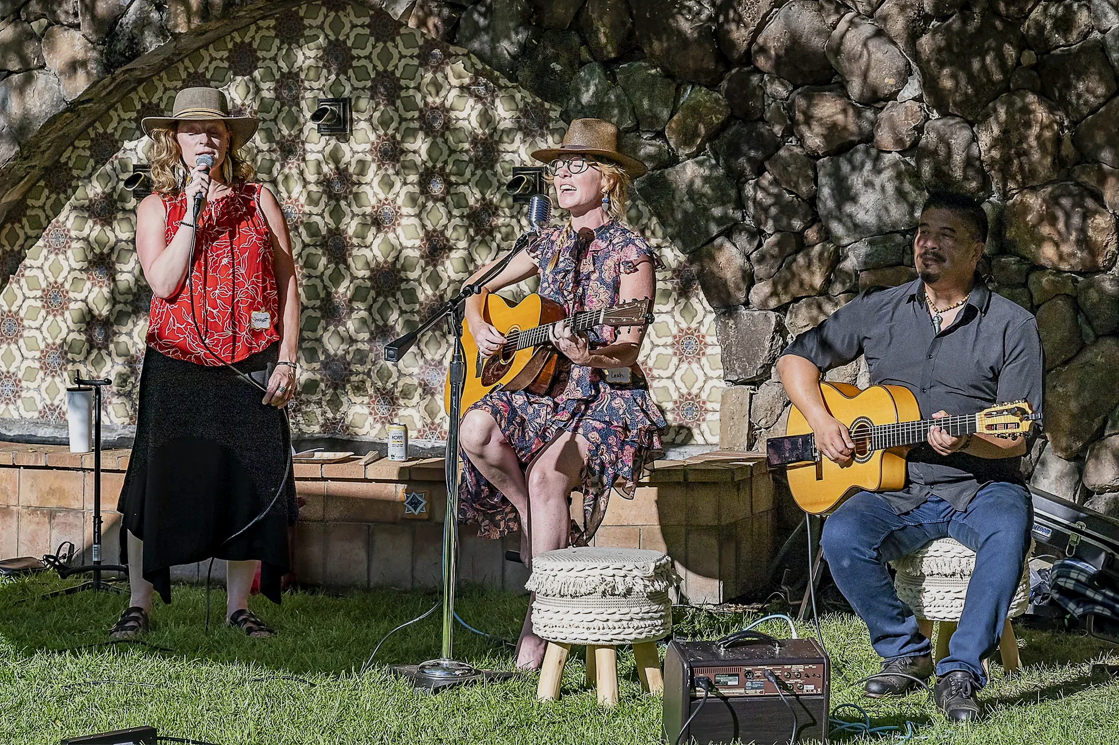Three musicians performing outdoors near a stone wall; one woman singing into a microphone, another woman playing guitar and singing, and a man playing guitar and listening.