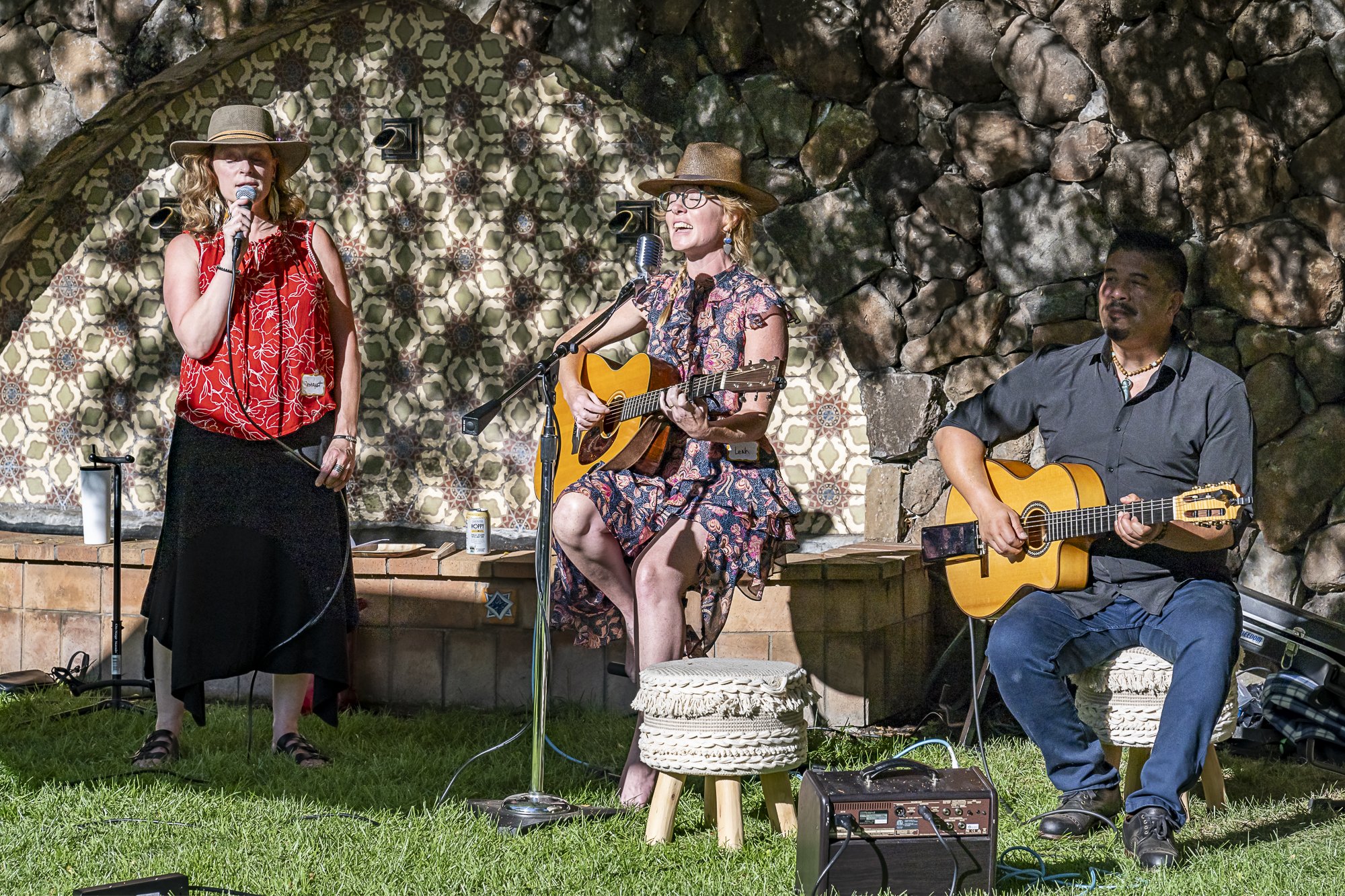 Three musicians performing outdoors in a rustic setting with stone walls. Two women with hats are singing, one playing an acoustic guitar, and a man with a guitar is seated on a stool. The scene appears casual and relaxed.