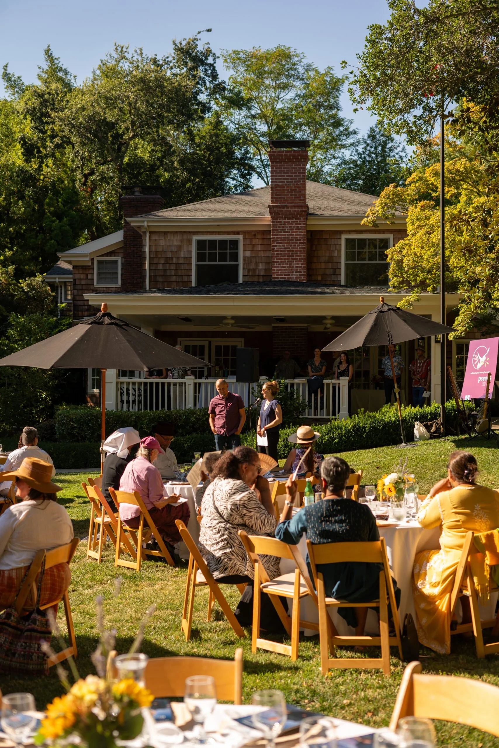 Outdoor gathering with people seated around tables on a lawn, in front of a house with a porch and a brick chimney, under large umbrellas, during daytime with trees and clear sky in the background.