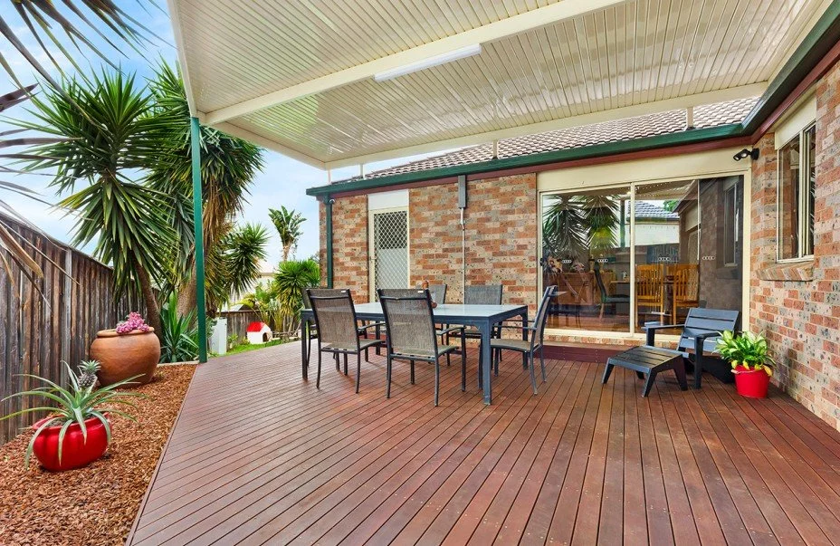 Outdoor wooden deck patio with a dining table and chairs, potted plants, and a view of a backyard with trees and a fence.