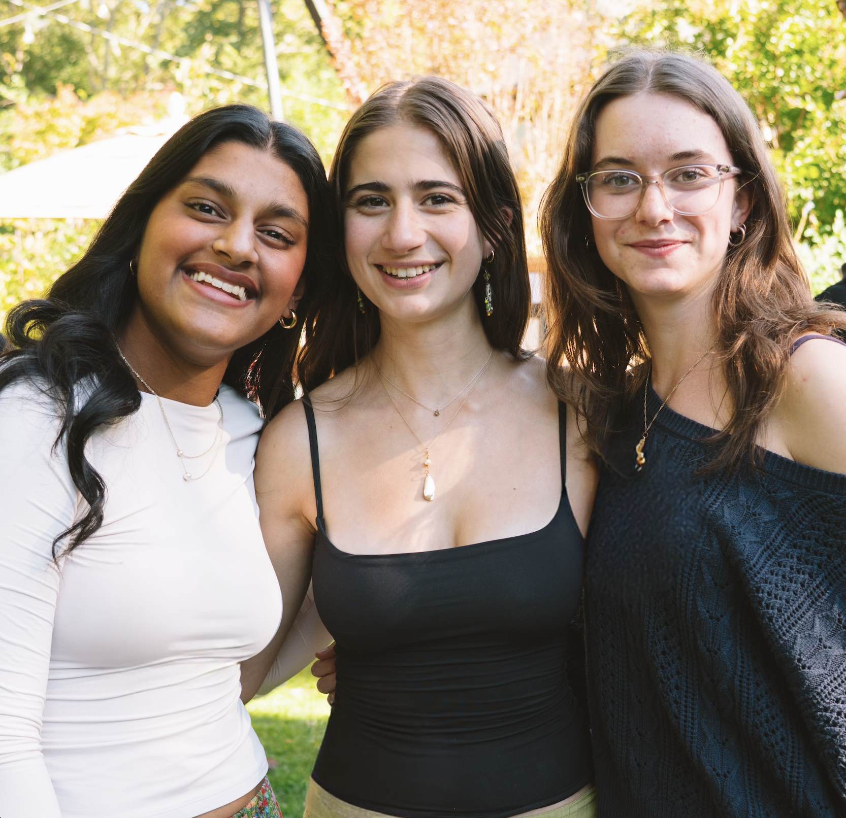 Three young women standing outdoors on a sunny day, smiling at the camera, with trees and greenery in the background.