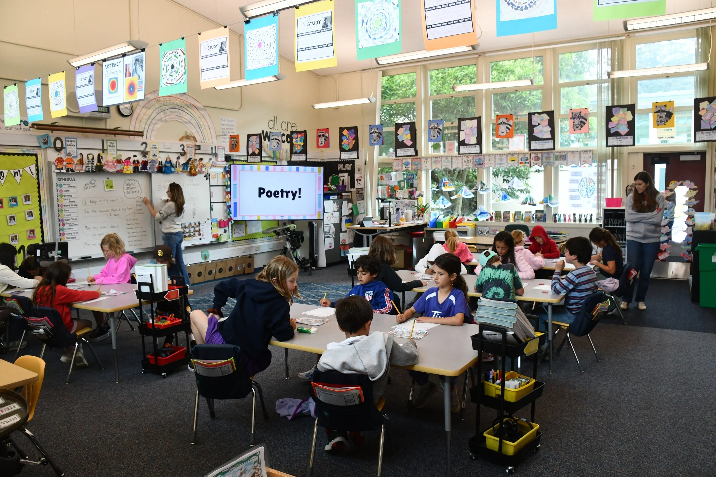 A classroom filled with students working at tables, with colorful student artwork hanging from the ceiling and walls, and a screen displaying the word 'Poetry!' at the front of the room.