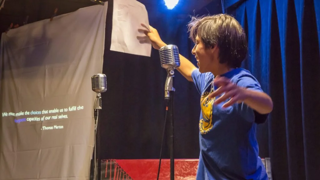 A person on stage gesturing with one hand while standing in front of a microphone, with another microphone nearby, and a quote projected on the curtain behind them.