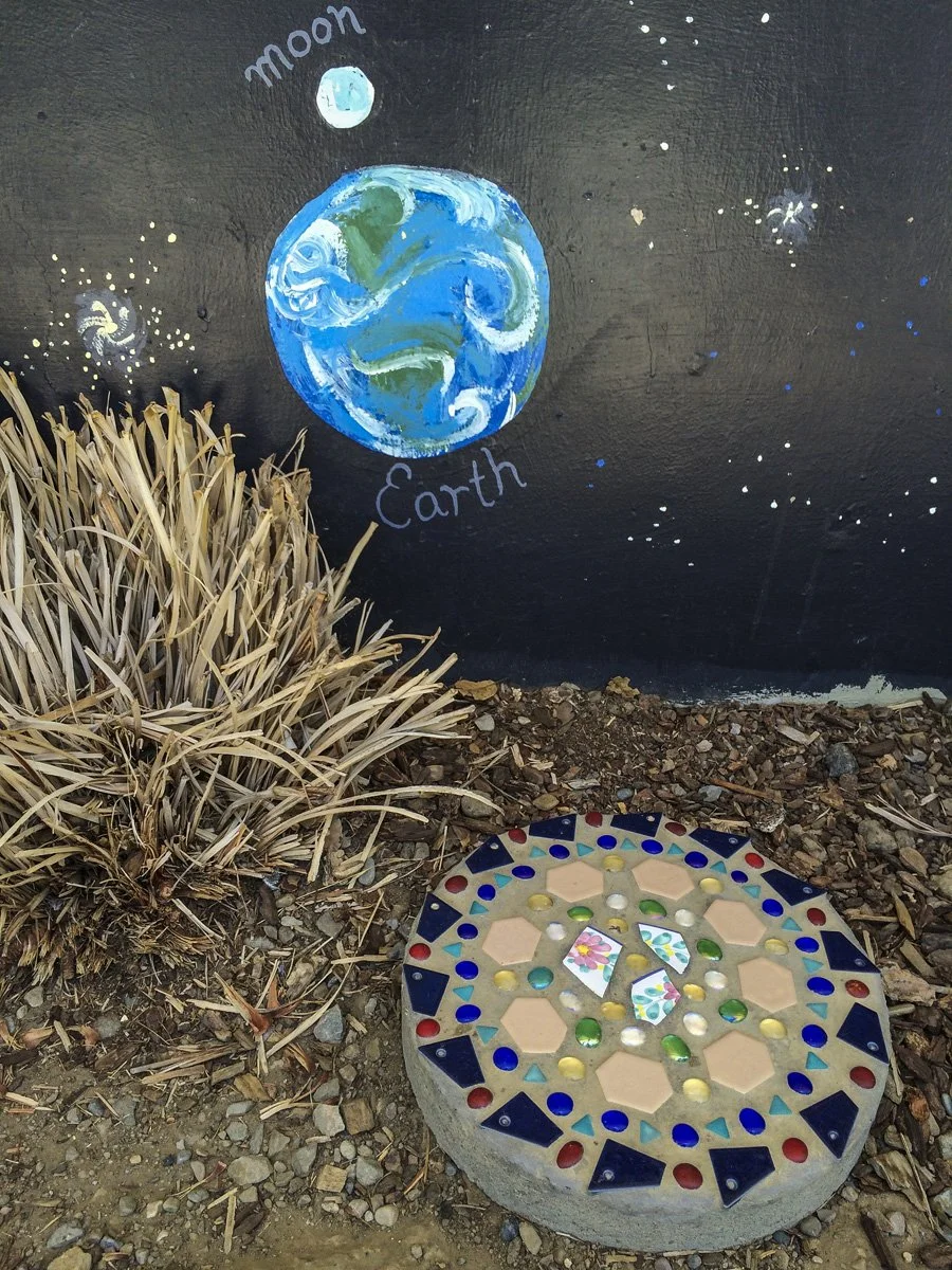 A decorative mosaic stone circle on the ground with colorful glass pieces, placed next to a dry bush. In the background, a painted mural of Earth on a black wall with the words 'Earth' and 'Moon,' and stars painted around.