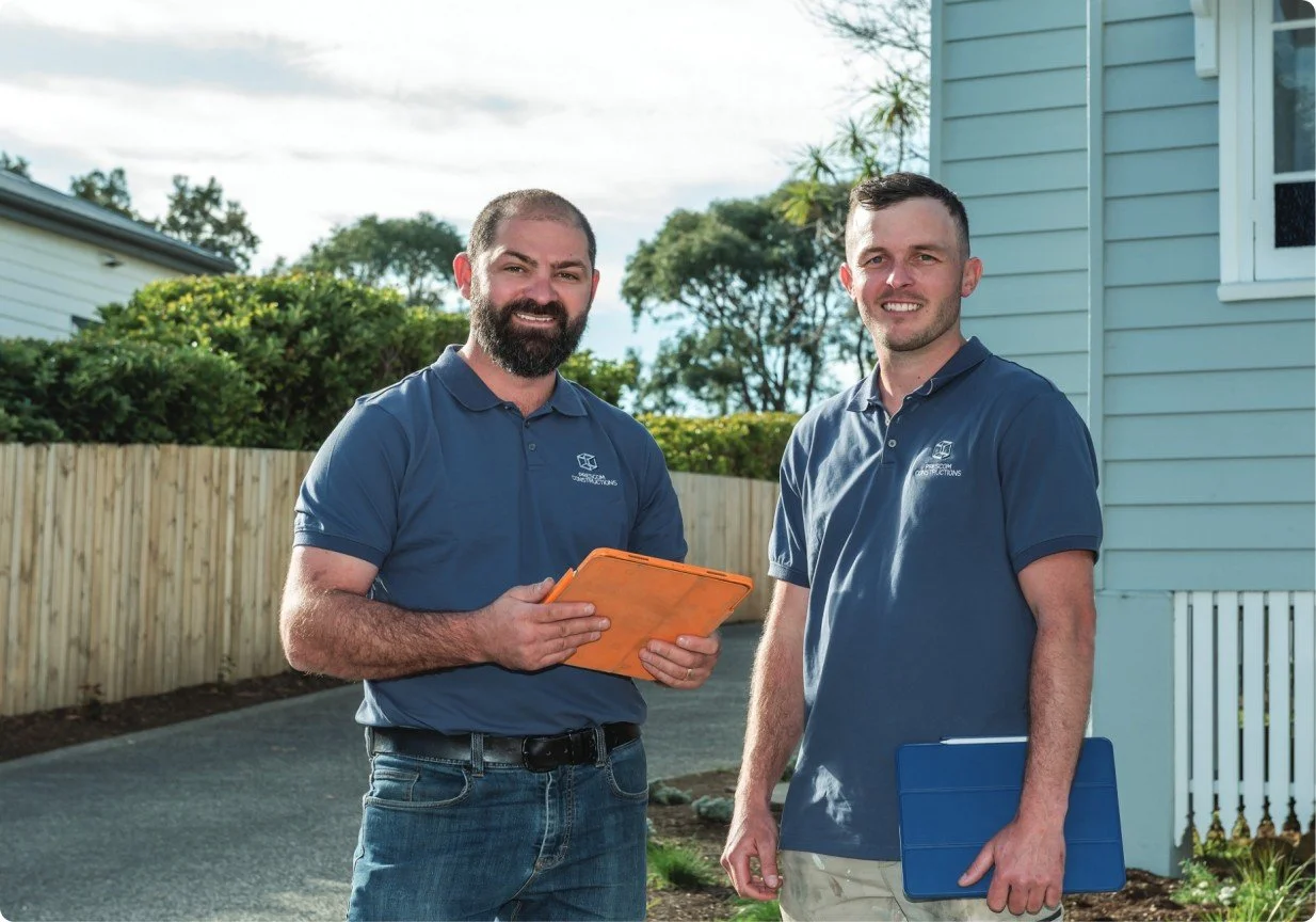 Two men standing outside near a house, wearing blue polo shirts with company logos, holding clipboards, smiling at the camera.