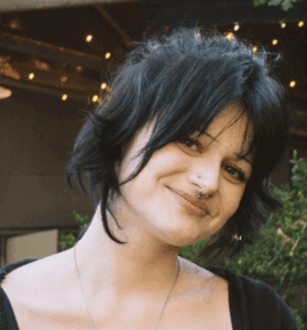 Close-up of a woman with black hair, septum piercing, and a smile, outdoors with fairy lights in the background.