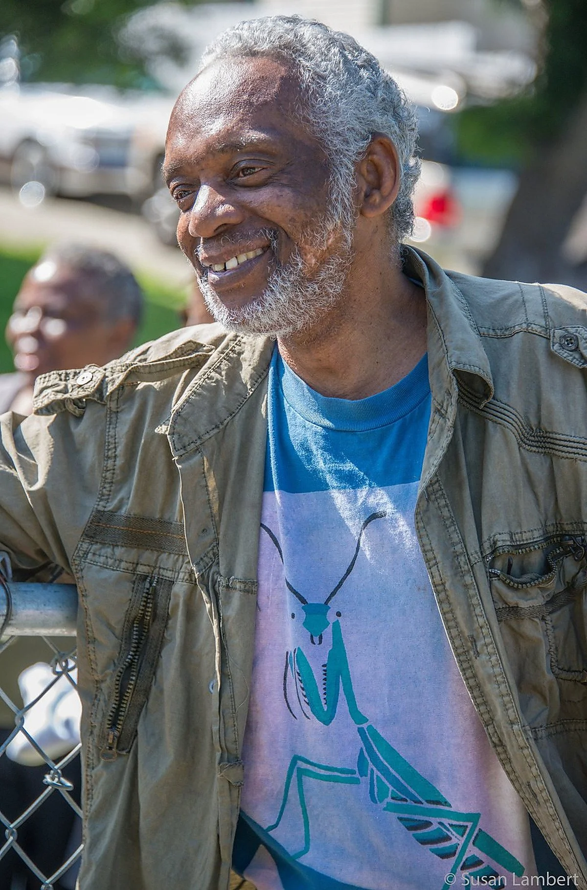 An elderly man with curly gray hair and a beard smiling, wearing a beige jacket over a graphic T-shirt, outdoors with blurred background of other people and cars.