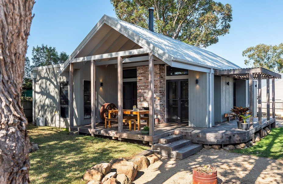 A modern house with a wooden deck, surrounded by trees and grass, featuring a sloped metal roof and a patio with outdoor furniture.