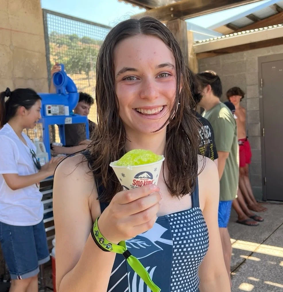 A smiling young woman with wet hair holding a cup of light green shaved ice, wearing a black sleeveless top and a neon green wristband, standing outdoors near a vending machine with other people in the background.