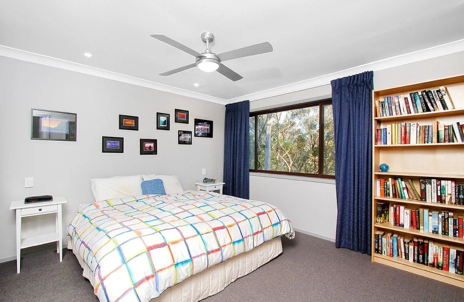 A bedroom with a bed, nightstands, a window with blue curtains, a ceiling fan, framed pictures on the wall, and a bookshelf filled with books.