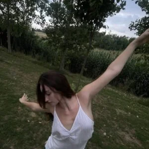 A woman with shoulder-length dark hair wearing a white sleeveless top outdoors, stretching her arms with a grassy area and trees in the background.