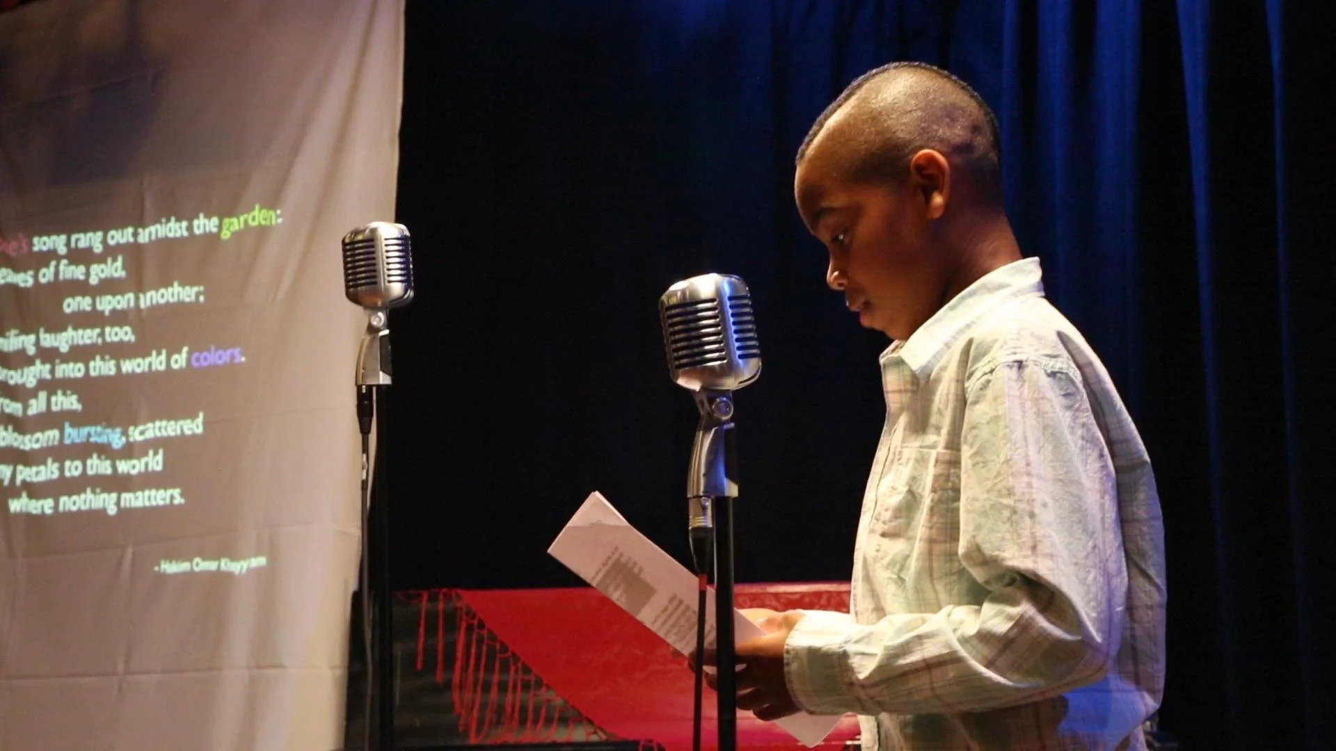 A young boy with a shaved head and a light-colored shirt standing at a red podium, reading from a piece of paper into a vintage silver microphone on a stage with dark blue curtains. To the left, a projection screen displays a poem with words highlighted in green, yellow, and purple.