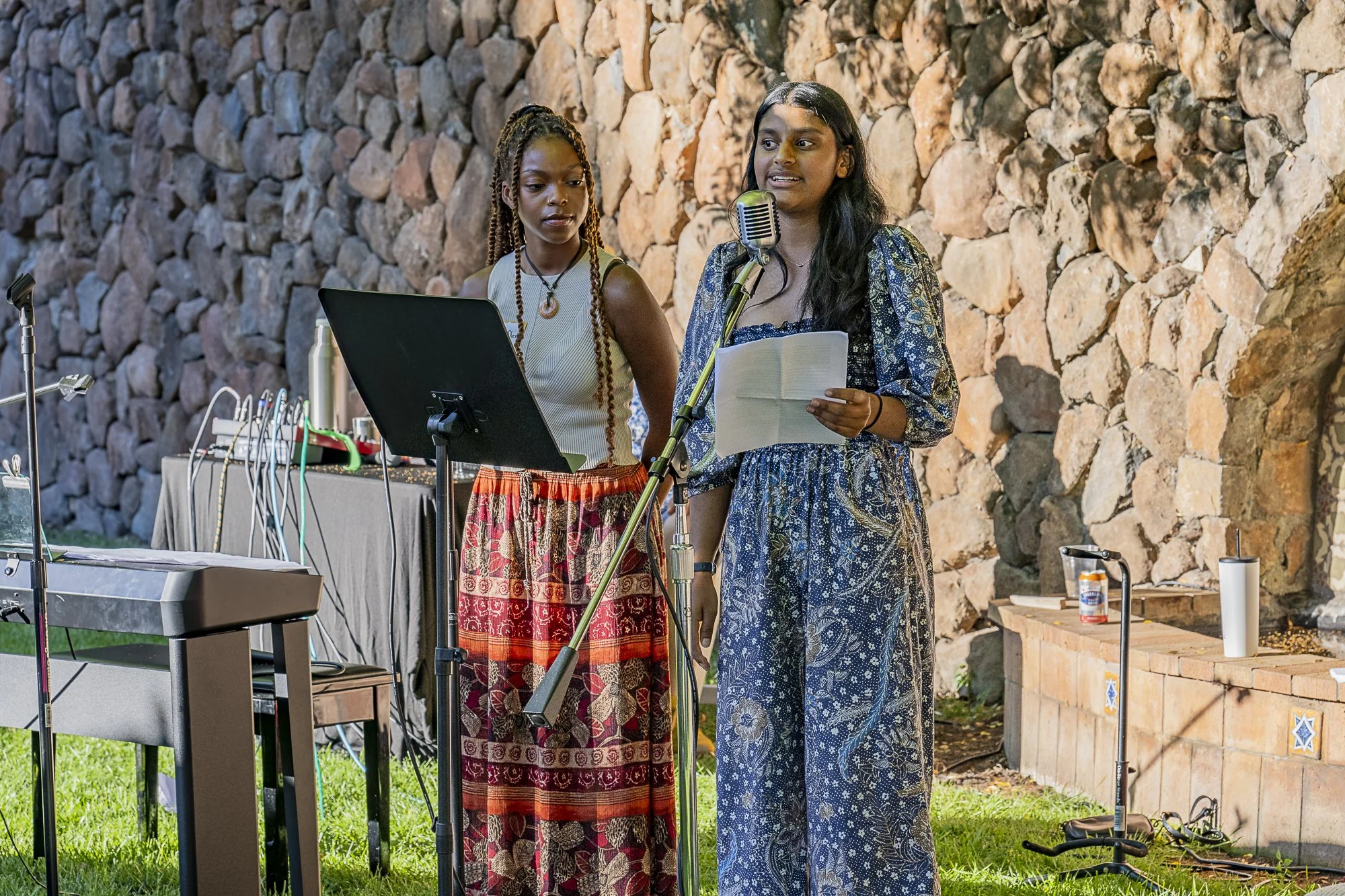 Two women speaking into a microphone outdoors with a rock wall in the background. One woman is holding a piece of paper, and the other is standing next to her. There is electronic equipment and a water bottle on a table nearby.
