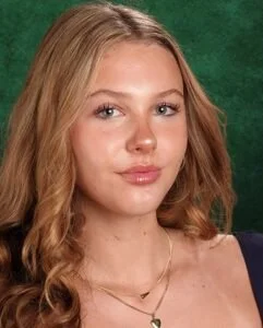 A young woman with long wavy blonde hair, wearing a necklace and a sleeveless top, standing against a green background.