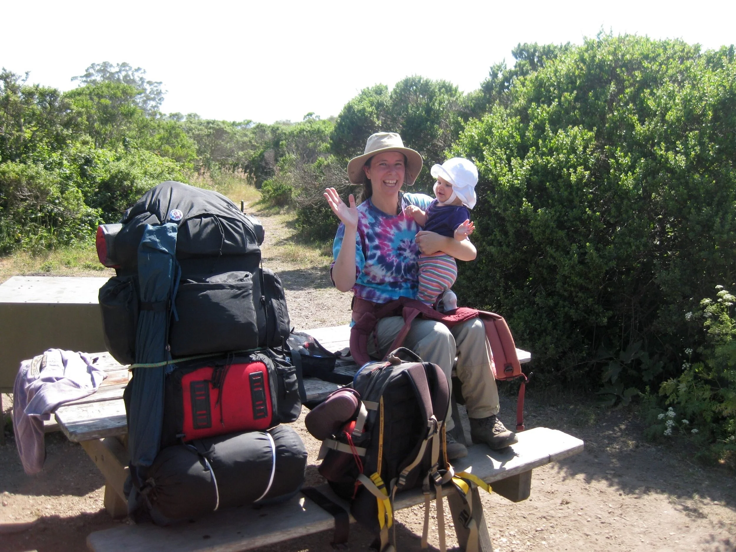 A woman smiling and holding a young girl on a park bench surrounded by a green natural landscape with backpacks and hiking gear nearby.