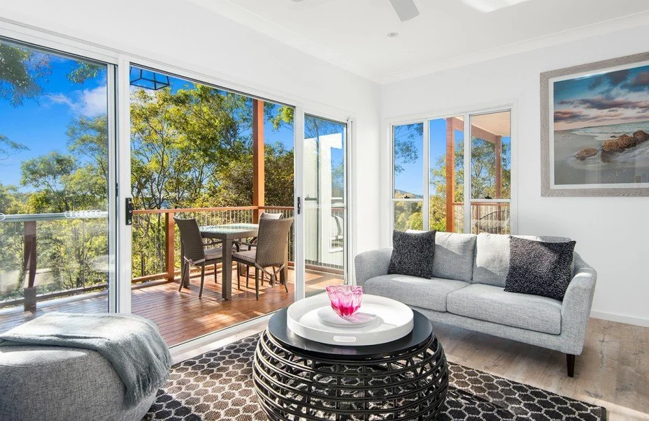Living room with large glass sliding doors leading to a balcony with outdoor furniture, a gray sofa with black cushions, a round black coffee table with a pink glass vase, and a mountain landscape painting on the wall.