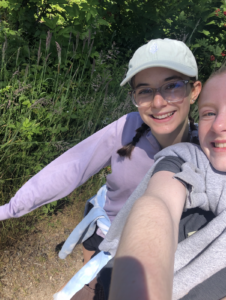 Two people taking a selfie outdoors with greenery in the background, one wearing a white cap and glasses, smiling.