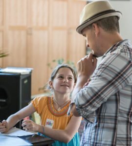 A man and a young girl sitting at a desk, smiling at each other. The man is wearing a hat and glasses, and the girl is wearing a yellow dress with beads.