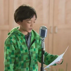 Young boy in a green checkered hoodie reading from a sheet of paper in front of a vintage microphone.