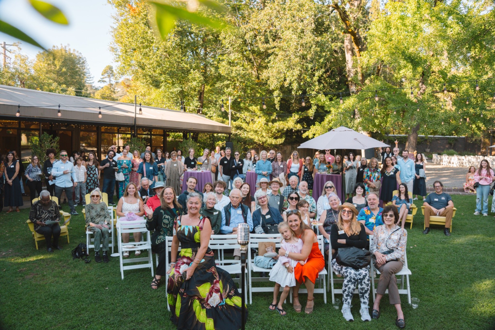 Group of diverse people gathered outdoors at what appears to be a community event, smiling and posing for a photo, with greenery and trees in the background.