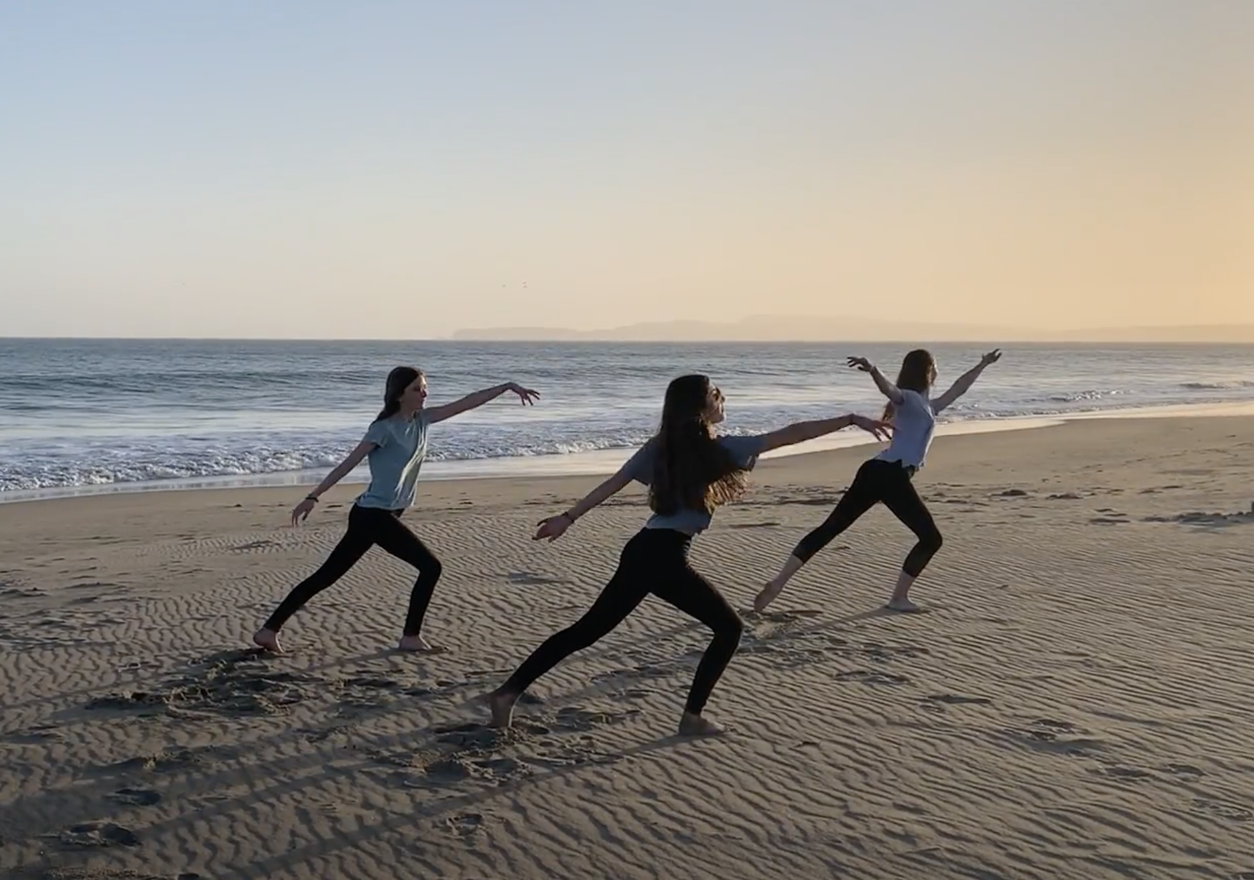 Three women practicing yoga on the beach at sunset, in warrior pose with the ocean in the background.