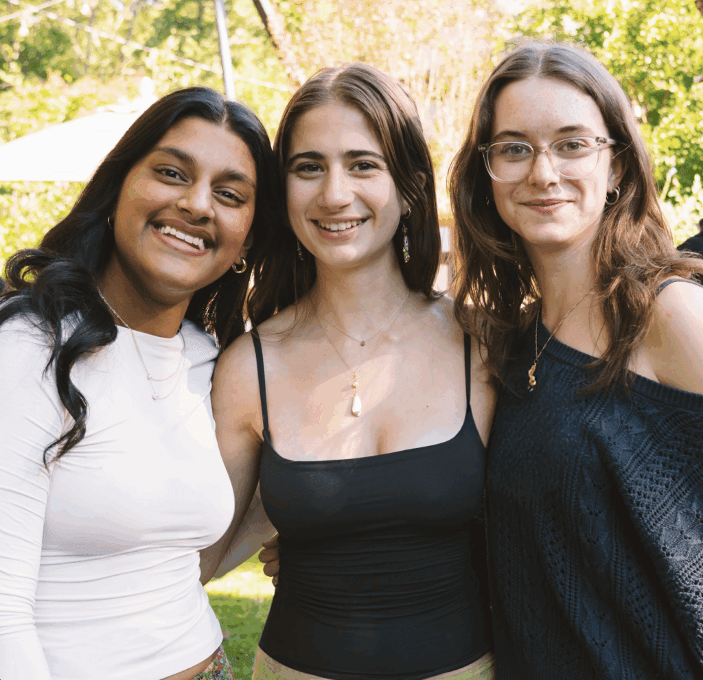 Three women smiling outdoors on a sunny day, standing close together with trees in the background.