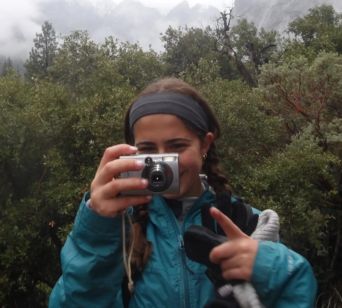 A girl with braided hair, wearing a blue jacket and a headband, taking a photo with a small silver camera in a foggy, wooded outdoor area.