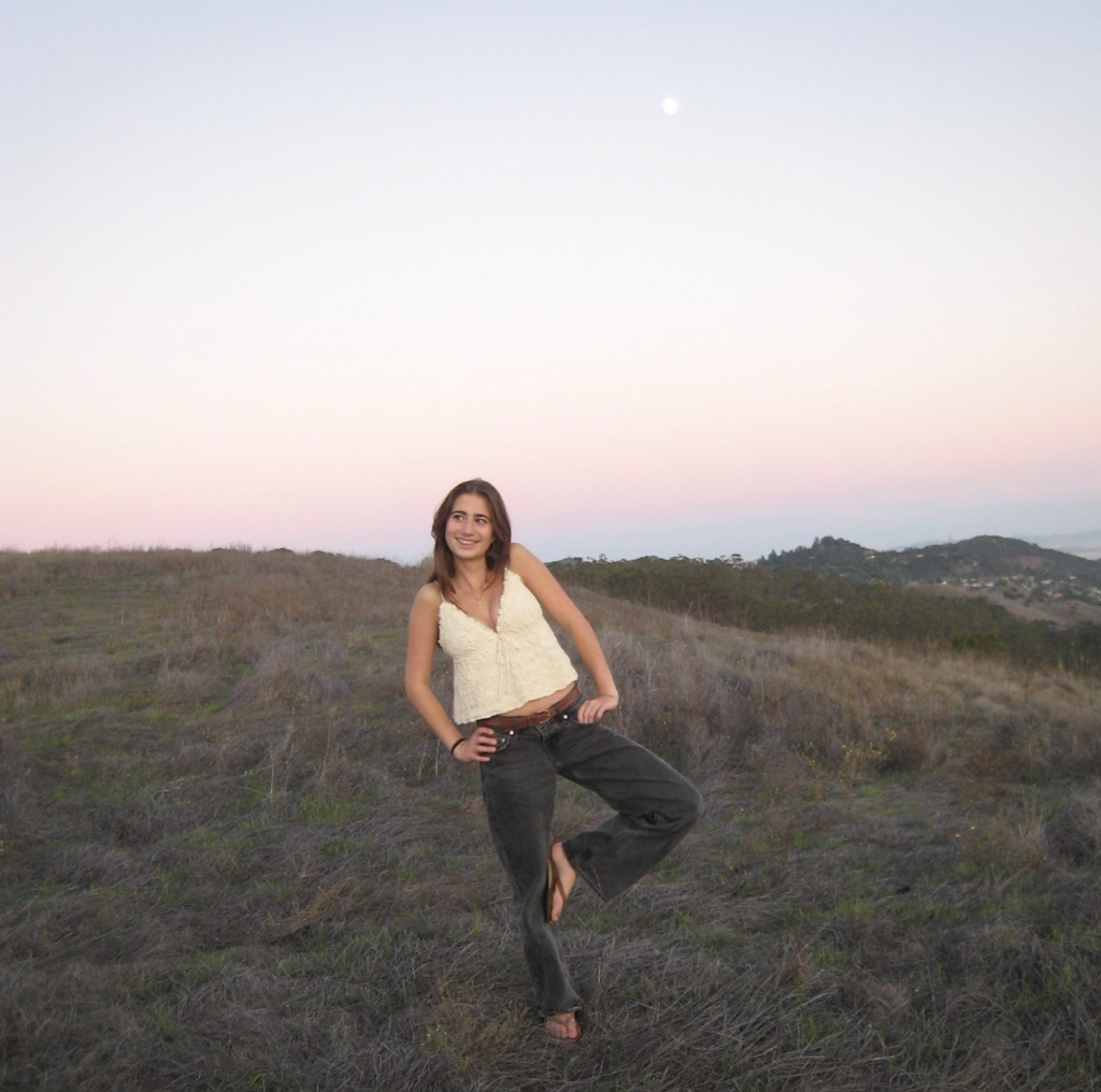 A young woman standing on a grassy hillside during sunset, wearing a cream-colored top and black pants, smiling with hand on hip and one leg bent.