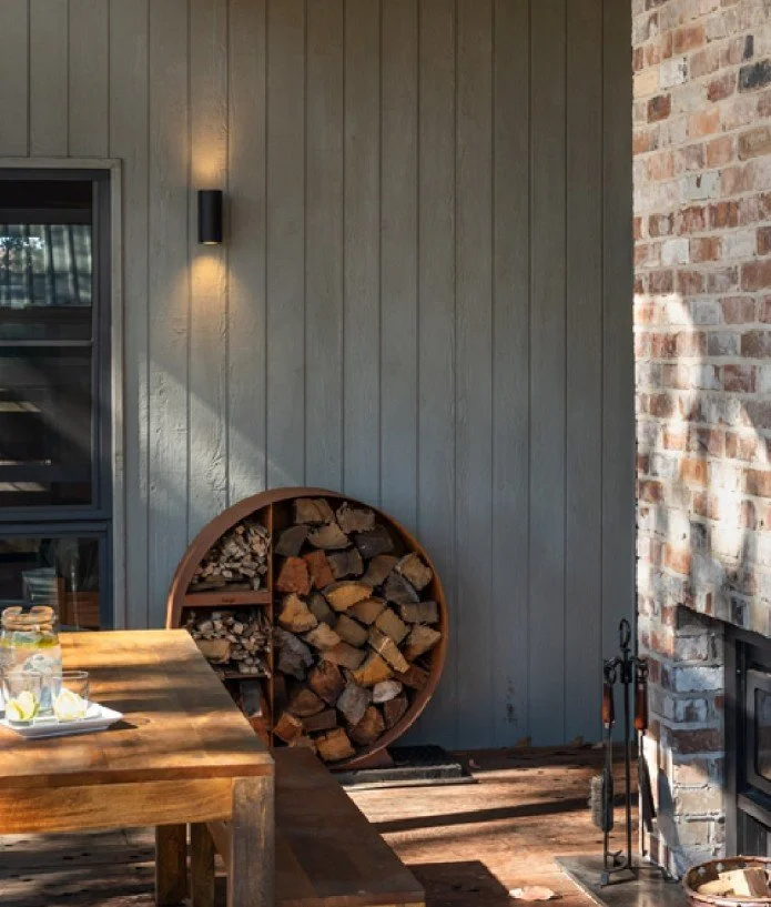 A backyard patio with a wood-fired fireplace, a round wood storage rack, a wooden table with drinks and snacks, and a wall-mounted light fixture.