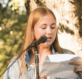 Young woman reading from a paper into a microphone outdoors with trees in the background.