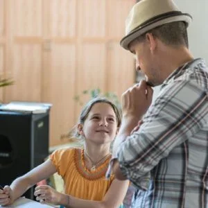 A man wearing a white hat and plaid shirt interacts with a young girl in an indoor setting, both smiling.
