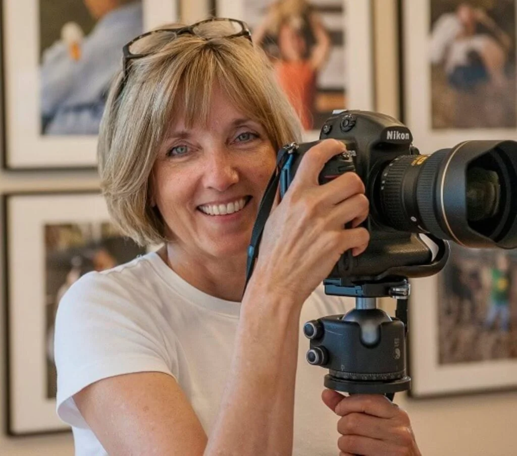 Woman with short blonde hair smiling while holding a Nikon camera with a long lens, standing in front of photos hanging on a wall.