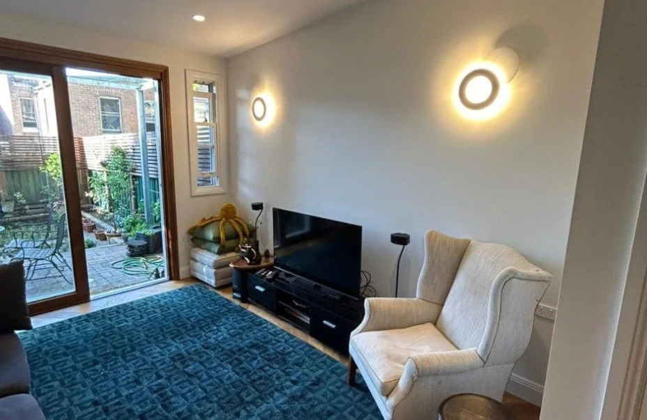 Living room with a sliding glass door leading to a backyard, a white armchair, a black TV on a stand, and two illuminated circular wall lights.